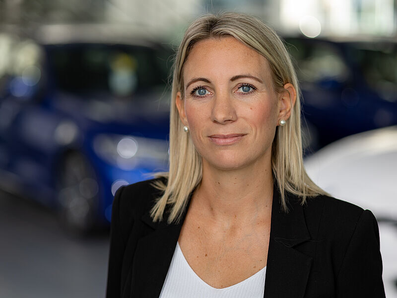 Kaltenbach Gruppe – Eine Frau mit glattem blondem Haar und blauen Augen, die einen schwarzen Blazer und ein weißes Oberteil trägt, steht in einem Innenraum in Kaltenbach-Remscheid mit verschwommenen blauen Autos im Hintergrund. Sie schaut mit neutralem Blick in die Kamera. – BMW, MINI, KIA, BMW Motorrad & Honda Motorrad Partner in NRW