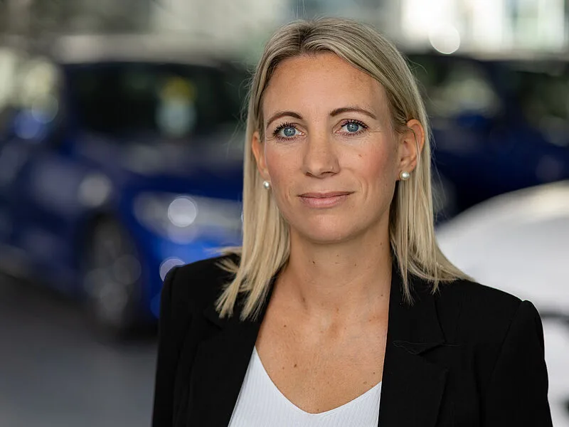 Kaltenbach Gruppe – Eine Frau mit glattem blondem Haar und blauen Augen, die einen schwarzen Blazer und ein weißes Oberteil trägt, steht in einem Innenraum in Kaltenbach-Remscheid mit verschwommenen blauen Autos im Hintergrund. Sie schaut mit neutralem Blick in die Kamera. – BMW, MINI, KIA, BMW Motorrad & Honda Motorrad Partner in NRW