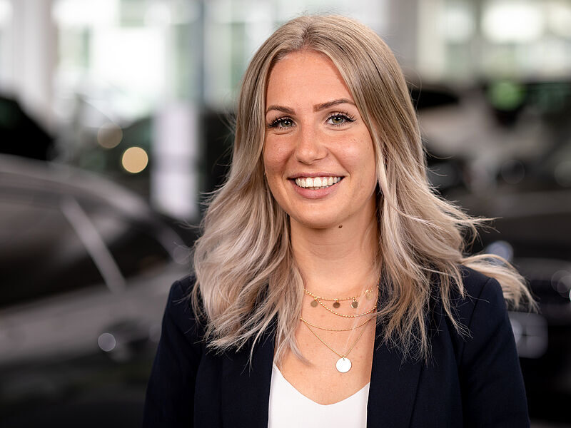 Kaltenbach Gruppe – Eine junge Frau mit langem, gewelltem blondem Haar lächelt in der Kaltenbach Zentrale in die Kamera. Sie trägt einen dunklen Blazer, ein weißes Oberteil und mehrere goldene Halsketten. Sie steht in einer hell erleuchteten Halle mit unscharfen Autos im Hintergrund. – BMW, MINI, KIA, BMW Motorrad & Honda Motorrad Partner in NRW