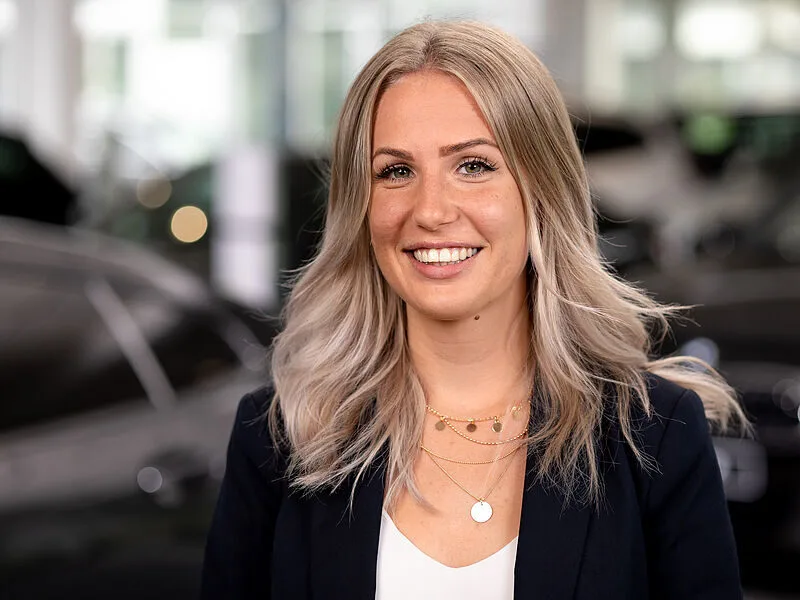 Kaltenbach Gruppe – Eine junge Frau mit langem, gewelltem blondem Haar lächelt in der Kaltenbach Zentrale in die Kamera. Sie trägt einen dunklen Blazer, ein weißes Oberteil und mehrere goldene Halsketten. Sie steht in einer hell erleuchteten Halle mit unscharfen Autos im Hintergrund. – BMW, MINI, KIA, BMW Motorrad & Honda Motorrad Partner in NRW