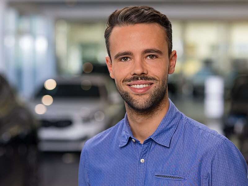 Kaltenbach Gruppe – Ein junger Mann mit kurzen braunen Haaren und gestutztem Bart, der ein blaues Button-up-Hemd trägt, lächelt in die Kamera. Der unscharfe Hintergrund deutet auf einen Innenbereich hin, möglicherweise Kaltenbach Overath, in dem Fahrzeuge zu sehen sind. – BMW, MINI, KIA, BMW Motorrad & Honda Motorrad Partner in NRW