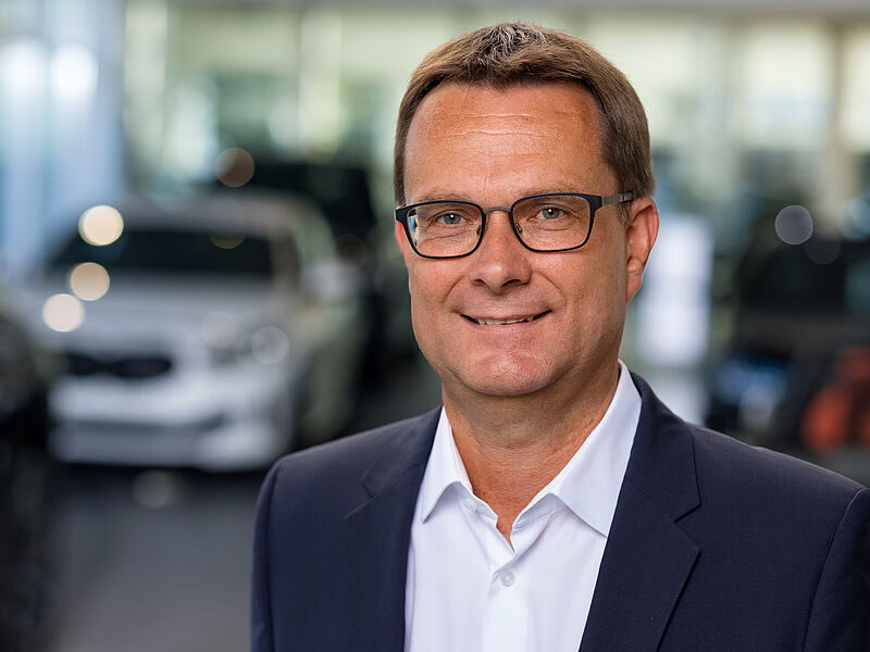 Kaltenbach Gruppe – Ein Mann mittleren Alters, der eine Brille, ein weißes Hemd und eine dunkle Anzugsjacke trägt, lächelt in einem Innenraum der Kaltenbach-Zentrale, während im Hintergrund unscharfe Autos zu sehen sind. – BMW, MINI, KIA, BMW Motorrad & Honda Motorrad Partner in NRW