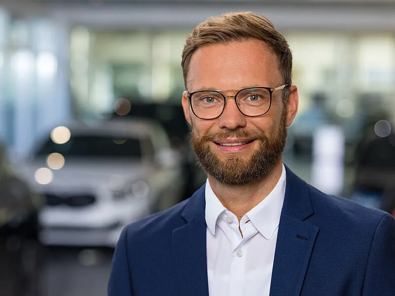Kaltenbach Gruppe – Ein lächelnder Mann mit Brille, Bart und kurzen Haaren, der eine blaue Anzugsjacke und ein weißes Hemd trägt, steht in einem Haus in Kaltenbach-Wiehl, mit unscharfen Autos im Hintergrund. – BMW, MINI, KIA, BMW Motorrad & Honda Motorrad Partner in NRW