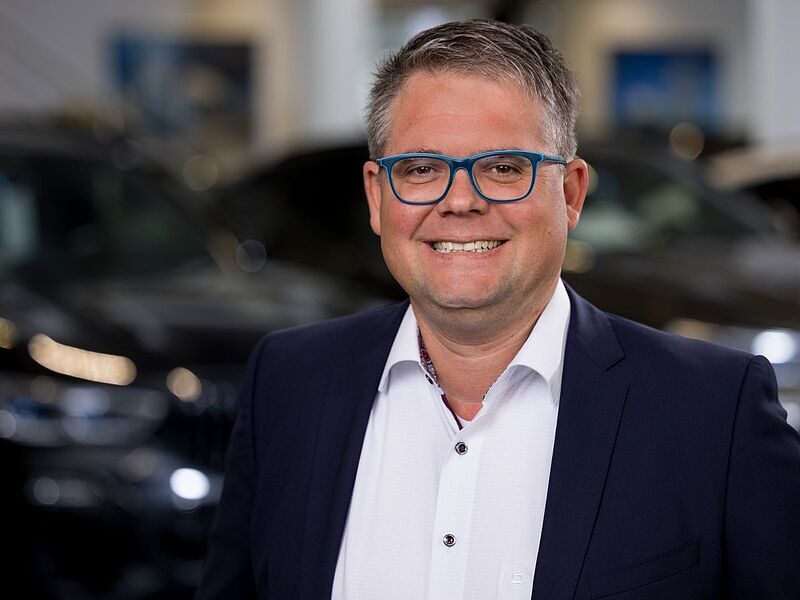 Kaltenbach Gruppe – Ein lächelnder Mann mit kurzen grauen Haaren, der eine blaue Brille, ein weißes Hemd und eine dunkle Anzugsjacke trägt, steht in einem Haus in Kaltenbach-Remscheid, mit verschwommenen Autos im Hintergrund. – BMW, MINI, KIA, BMW Motorrad & Honda Motorrad Partner in NRW