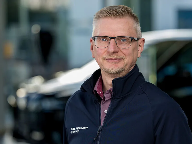 Kaltenbach Gruppe – Ein Mann mit kurzen blonden Haaren und Brille, der eine marineblaue Jacke mit der Aufschrift Kaltenbach Gruppe trägt, steht im Freien vor einem unscharfen Hintergrund mit einem dunklen Fahrzeug und einem Gebäude in Kaltenbach Wermelskirchen. – BMW, MINI, KIA, BMW Motorrad & Honda Motorrad Partner in NRW