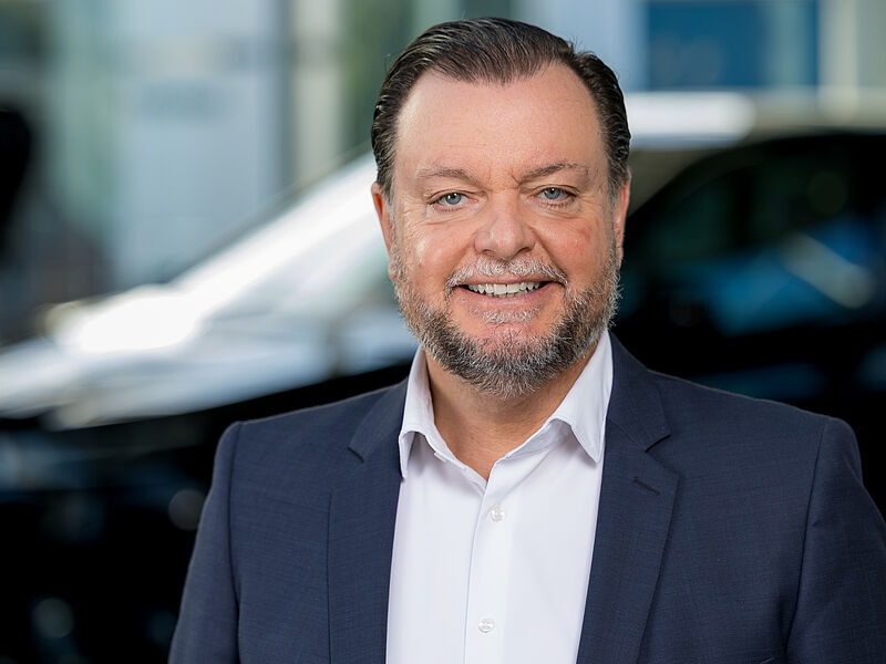 Kaltenbach Gruppe – Ein lächelnder Mann mit kurzen braunen Haaren und einem Bart, der eine marineblaue Anzugsjacke und ein weißes Hemd trägt, steht vor einem unscharfen Hintergrund mit einem Auto in Kaltenbach Wermelskirchen. – BMW, MINI, KIA, BMW Motorrad & Honda Motorrad Partner in NRW