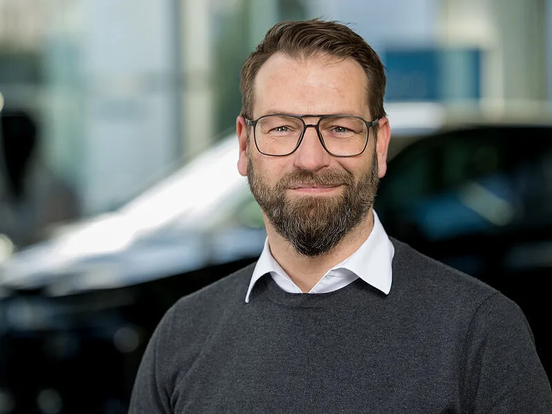 Kaltenbach Gruppe – Ein Mann mit Brille, Bart und Schnurrbart, der ein weißes Kragenhemd unter einem dunklen Pullover trägt, lächelt in die Kamera. Der unscharfe Hintergrund suggeriert einen Innenraum in Kaltenbach Wermelskirchen mit sanftem Tageslicht. – BMW, MINI, KIA, BMW Motorrad & Honda Motorrad Partner in NRW