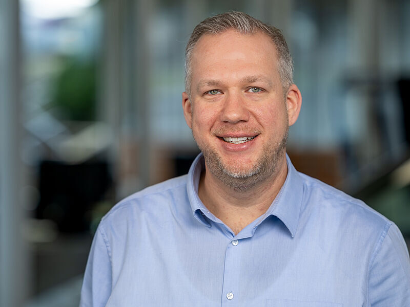Kaltenbach Gruppe – Ein lächelnder Mann mit kurzen hellen Haaren und gestutztem Bart, der ein hellblaues Button-up-Hemd trägt, steht in einem Innenraum mit einem unscharfen Hintergrund des Büros in Kaltenbach Wipperfürth. – BMW, MINI, KIA, BMW Motorrad & Honda Motorrad Partner in NRW