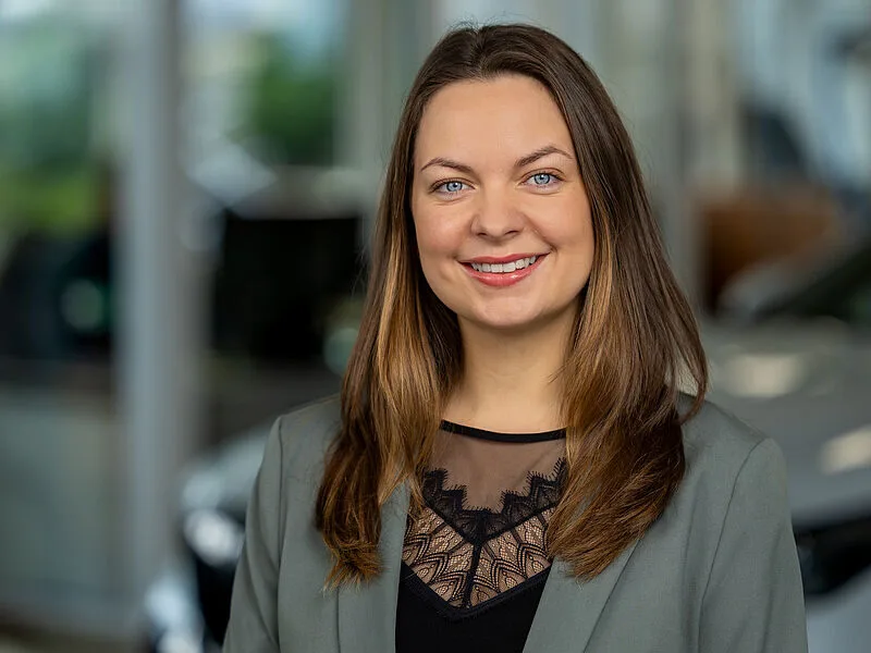 Kaltenbach Gruppe – Eine Frau mit langen braunen Haaren, die einen grauen Blazer über einem schwarzen Oberteil mit Spitzenausschnitt trägt, lächelt in einem Innenraum mit gedämpftem Licht in Kaltenbach Wipperfürth in die Kamera. Der Hintergrund ist unscharf. – BMW, MINI, KIA, BMW Motorrad & Honda Motorrad Partner in NRW