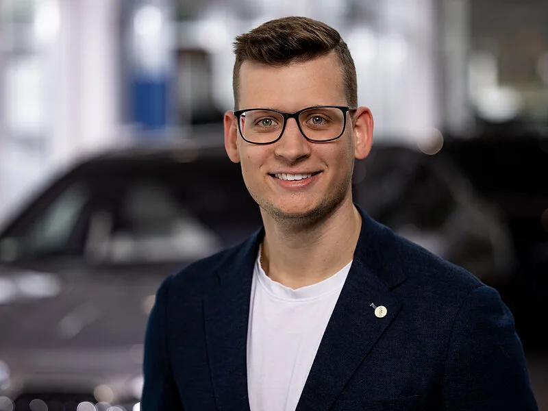 Kaltenbach Gruppe – Ein lächelnder Mann mit kurzen Haaren und Brille, der einen dunklen Blazer über einem weißen Hemd trägt, steht in einem Innenraum der Kaltenbach-Zentrale, mit verschwommenen Autos und einem hellen Hintergrund hinter ihm. – BMW, MINI, KIA, BMW Motorrad & Honda Motorrad Partner in NRW