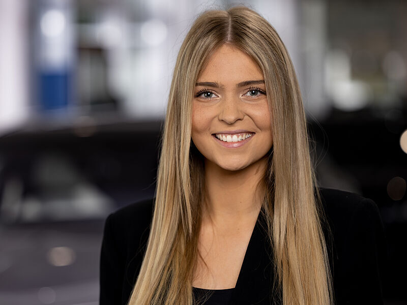 Kaltenbach Gruppe – Eine junge Frau mit langen glatten blonden Haaren lächelt in der Kaltenbach Zentrale in die Kamera. Sie trägt ein schwarzes Oberteil und einen Blazer und hat einen unscharfen Innenhintergrund im Rücken. – BMW, MINI, KIA, BMW Motorrad & Honda Motorrad Partner in NRW