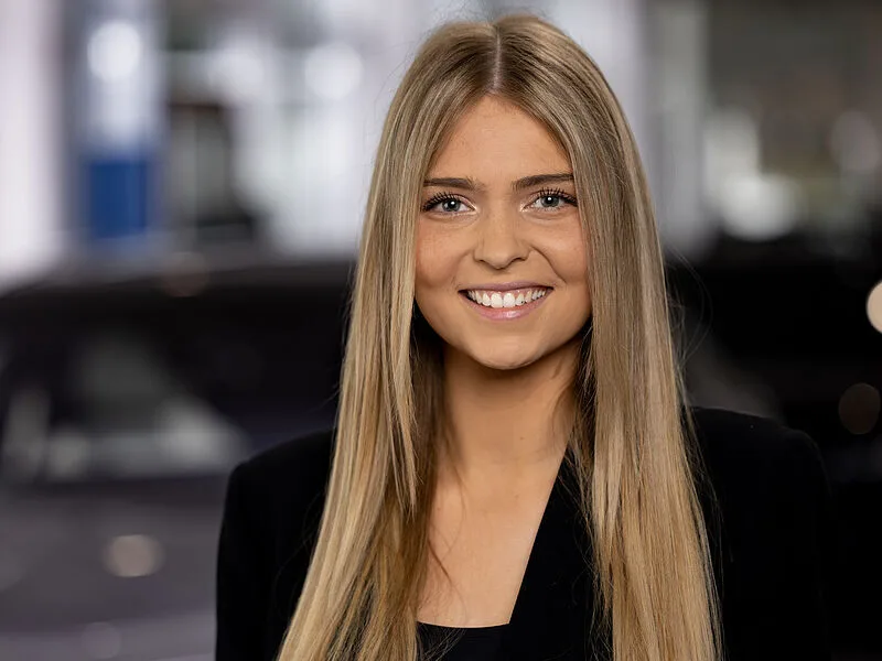 Kaltenbach Gruppe – Eine junge Frau mit langen glatten blonden Haaren lächelt in der Kaltenbach Zentrale in die Kamera. Sie trägt ein schwarzes Oberteil und einen Blazer und hat einen unscharfen Innenhintergrund im Rücken. – BMW, MINI, KIA, BMW Motorrad & Honda Motorrad Partner in NRW