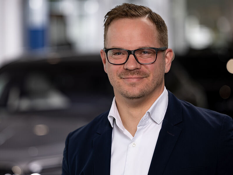 Kaltenbach Gruppe – Ein Mann mit kurzen hellbraunen Haaren, Brille und gestutztem Bart trägt einen dunklen Blazer über einem weißen Hemd und lächelt leicht, während er in einem Innenraum mit unscharfem Hintergrund im Büro von Kaltenbach Wiehl steht. – BMW, MINI, KIA, BMW Motorrad & Honda Motorrad Partner in NRW