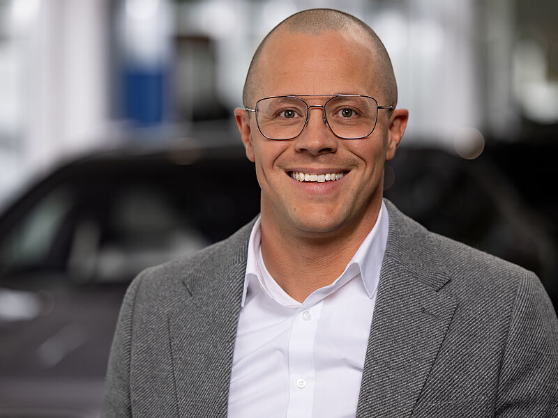 Kaltenbach Gruppe – Ein lächelnder Mann mit Brille, rasiertem Kopf und einem grauen Blazer über einem weißen Hemd steht in einem Innenraum der Kaltenbach-Zentrale, mit verschwommenen Autos und Lichtern im Hintergrund. – BMW, MINI, KIA, BMW Motorrad & Honda Motorrad Partner in NRW