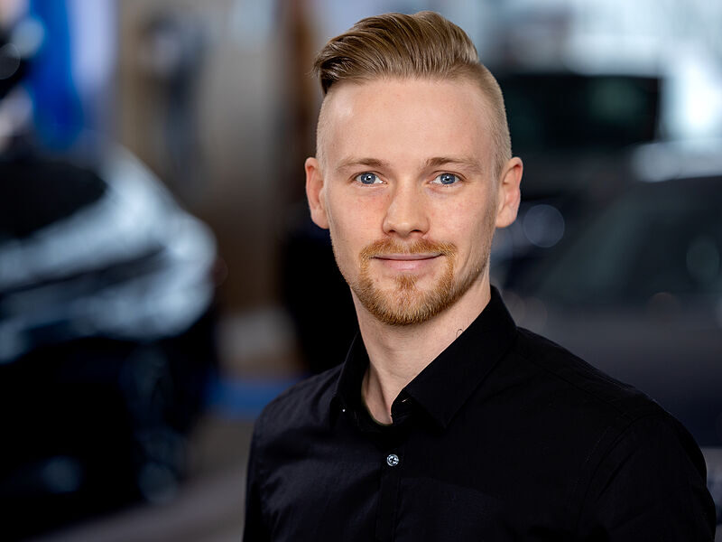 Kaltenbach Gruppe – Ein Mann mit kurzen, blonden, zur Seite gestylten Haaren und einem gestutzten Bart lächelt in die Kamera. Er trägt ein schwarzes Button-up-Hemd. Der unscharfe Hintergrund deutet auf einen Innenraum hin, möglicherweise auf eine Veranstaltung in Kaltenbach Olpe. – BMW, MINI, KIA, BMW Motorrad & Honda Motorrad Partner in NRW