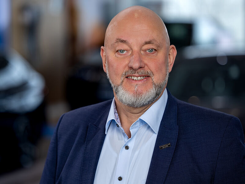 Kaltenbach Gruppe – Ein Mann mittleren Alters mit einer Glatze, grauem Bart und blauen Augen lächelt. Er trägt eine blaue Anzugjacke über einem hellblauen Hemd und steht in einem Raum, der die Kaltenbach-Zentrale zu sein scheint, wobei der Hintergrund in Innenräumen leicht verschwommen ist. – BMW, MINI, KIA, BMW Motorrad & Honda Motorrad Partner in NRW