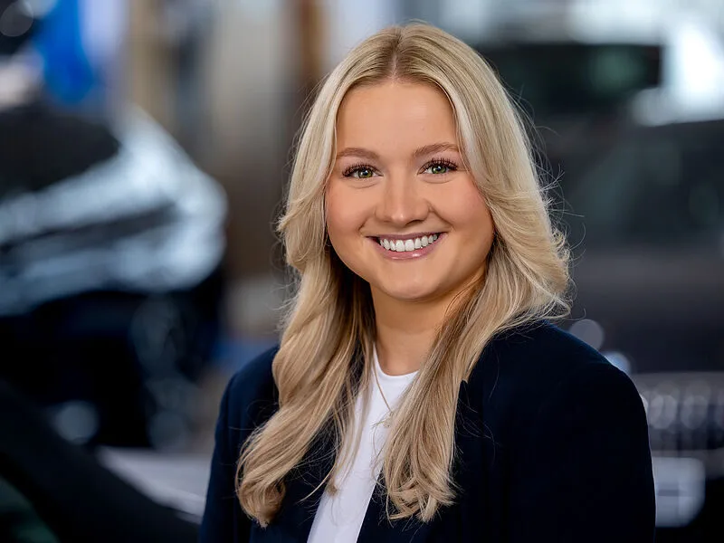 Kaltenbach Gruppe – Eine junge Frau mit langen blonden Haaren, die einen dunklen Blazer und ein weißes Oberteil trägt, lächelt in einem hellen, unscharfen Innenraum, möglicherweise einem Autohaus in Kaltenbach Waldbröl, in die Kamera. – BMW, MINI, KIA, BMW Motorrad & Honda Motorrad Partner in NRW