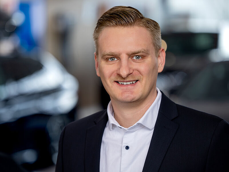 Kaltenbach Gruppe – Ein lächelnder Mann mit kurzen blonden Haaren, dunkler Anzugjacke und weißem Button-up-Hemd steht in einem Innenraum der Kaltenbach-Zentrale, mit verschwommenen Autos und Lichtern eines Ausstellungsraums im Hintergrund. – BMW, MINI, KIA, BMW Motorrad & Honda Motorrad Partner in NRW