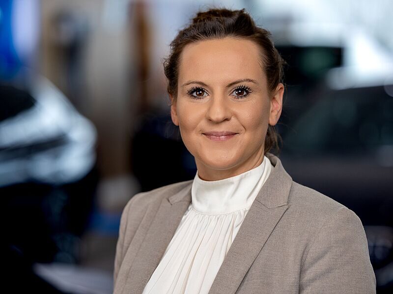 Kaltenbach Gruppe – Eine Frau mit zurückgebundenem braunem Haar, die einen hellgrauen Blazer und eine weiße hochgeschlossene Bluse trägt, lächelt in die Kamera. Der unscharfe Hintergrund deutet auf einen Innenraum in Kaltenbach Olpe hin. – BMW, MINI, KIA, BMW Motorrad & Honda Motorrad Partner in NRW