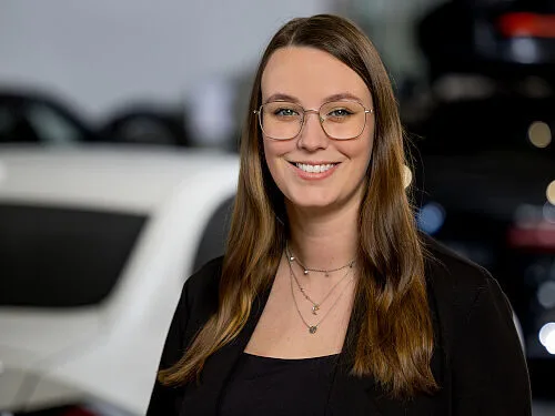 Kaltenbach Gruppe – Eine Frau mit langen braunen Haaren und Brille lächelt in die Kamera. Sie trägt ein schwarzes Oberteil, einen schwarzen Blazer und mehrlagige Ketten. Im Hintergrund sind unscharfe Autos in Kaltenbach Meinerzhagen zu sehen. – BMW, MINI, KIA, BMW Motorrad & Honda Motorrad Partner in NRW