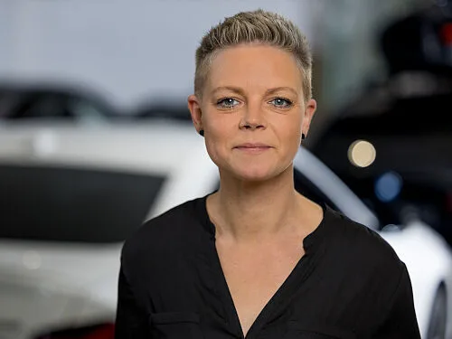 Kaltenbach Gruppe – Eine Person mit kurzen, hellen Haaren und schwarzem Hemd steht vor einem unscharfen Hintergrund mit weißen und dunkelfarbigen Autos in Kaltenbach Meinerzhagen, schaut in die Kamera und lächelt leicht. – BMW, MINI, KIA, BMW Motorrad & Honda Motorrad Partner in NRW