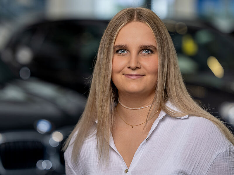 Kaltenbach Gruppe – Eine junge Frau mit langen blonden Haaren und blauen Augen, die eine weiße Bluse und eine zarte Halskette trägt, lächelt sanft, während sie in einem Innenraum vor einem verschwommenen dunklen Auto in Kaltenbach Remscheid steht. – BMW, MINI, KIA, BMW Motorrad & Honda Motorrad Partner in NRW