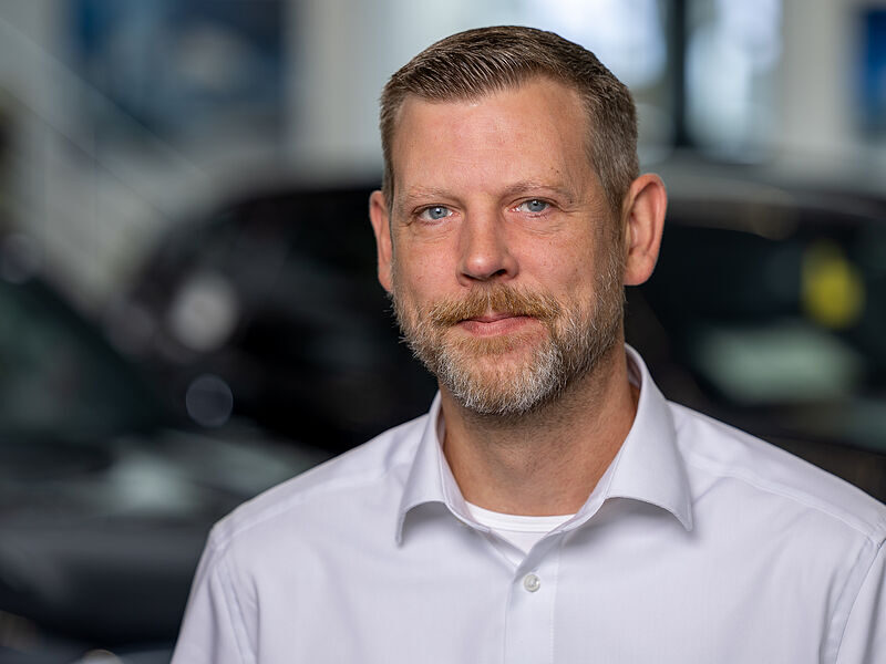 Kaltenbach Gruppe – Ein Mann mit kurzen hellbraunen Haaren, einem gestutzten Bart und einem Schnurrbart, der ein weißes Hemd mit Kragen trägt, steht in einem Haus in Kaltenbach-Remscheid mit unscharfen Autos im Hintergrund. – BMW, MINI, KIA, BMW Motorrad & Honda Motorrad Partner in NRW