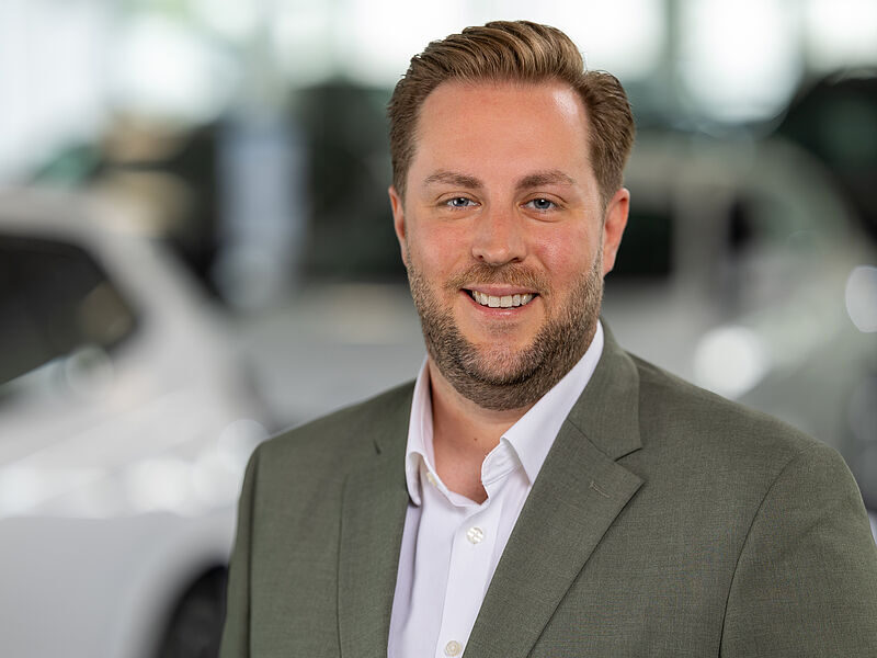 Kaltenbach Gruppe – Ein lächelnder Mann mit kurzen hellbraunen Haaren und Bart, der einen grauen Blazer und ein weißes Hemd trägt, steht in einem Haus in Kaltenbach-Wiehl, mit unscharfen Autos im Hintergrund. – BMW, MINI, KIA, BMW Motorrad & Honda Motorrad Partner in NRW