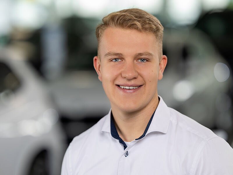 Kaltenbach Gruppe – Ein junger Mann mit kurzen blonden Haaren, der ein weißes Button-up-Hemd trägt, lächelt in die Kamera. Der unscharfe Hintergrund deutet auf einen Innenbereich hin, möglicherweise den Ausstellungsraum oder das Büro von Kaltenbach Overath. – BMW, MINI, KIA, BMW Motorrad & Honda Motorrad Partner in NRW