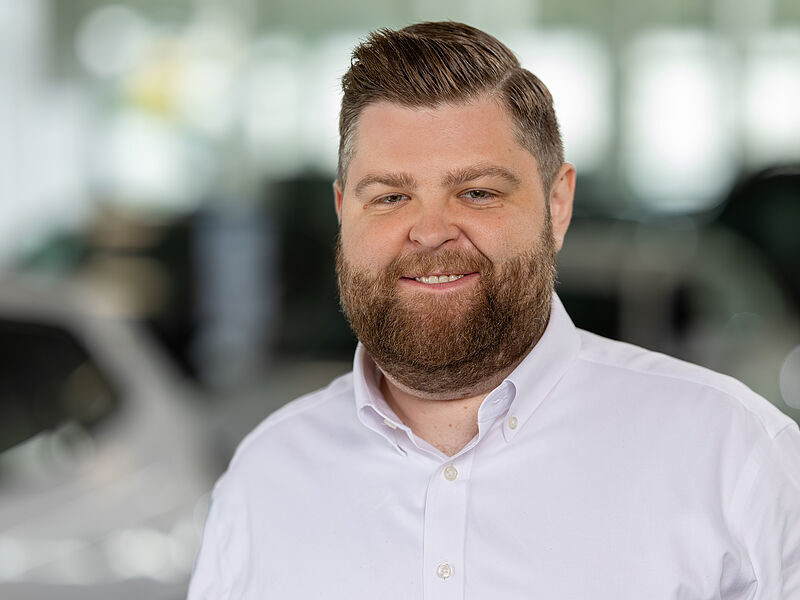 Kaltenbach Gruppe – Ein lächelnder Mann mit Bart und kurzen Haaren, der ein weißes Hemd mit Kragen trägt, steht in einem Haus in Kaltenbach Overath, mit verschwommenen Autos und hellen Fenstern im Hintergrund. – BMW, MINI, KIA, BMW Motorrad & Honda Motorrad Partner in NRW