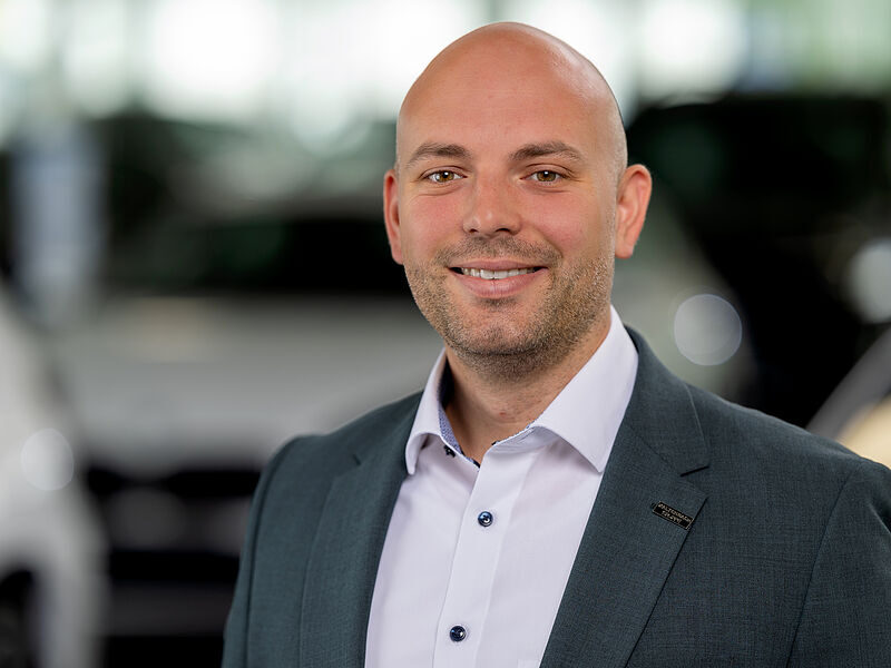 Kaltenbach Gruppe – Ein lächelnder, glatzköpfiger Mann in grauem Anzug und weißem Hemd steht in einem Innenraum der Kaltenbach Zentrale, im Hintergrund sind unscharfe Fahrzeuge zu sehen. – BMW, MINI, KIA, BMW Motorrad & Honda Motorrad Partner in NRW