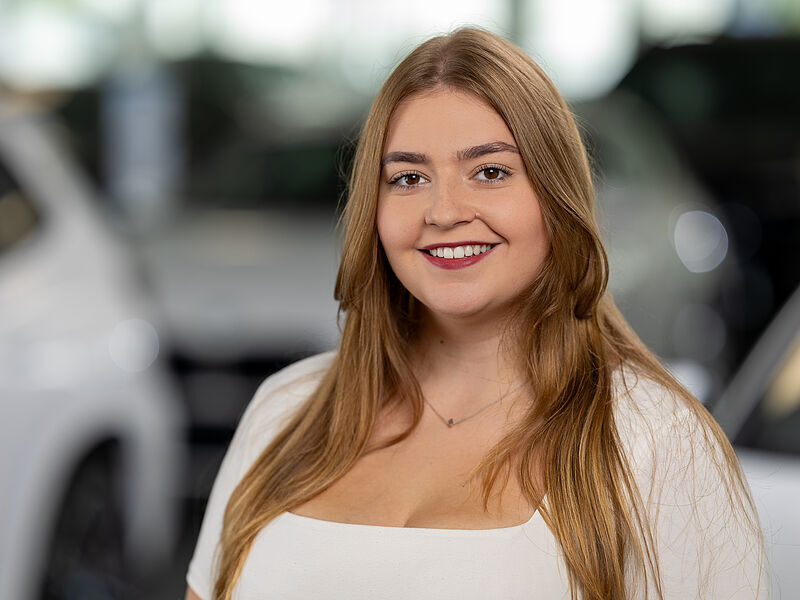 Kaltenbach Gruppe – Eine junge Frau mit langen, hellbraunen Haaren lächelt in Kaltenbach Waldbröl in die Kamera. Sie trägt ein weißes Oberteil und eine zarte Halskette. Im Hintergrund sind unscharfe weiße Autos zu sehen. – BMW, MINI, KIA, BMW Motorrad & Honda Motorrad Partner in NRW