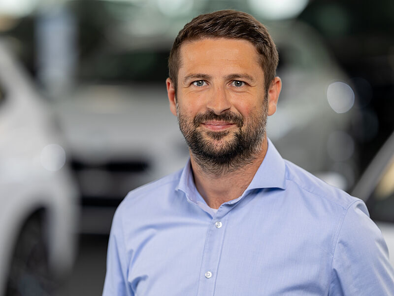 Kaltenbach Gruppe – Ein Mann mit kurzen braunen Haaren und einem Bart, der ein hellblaues Button-up-Hemd trägt, steht in einem Haus in Kaltenbach-Wiehl mit unscharfen Autos im Hintergrund. Er lächelt leicht und schaut in die Kamera. – BMW, MINI, KIA, BMW Motorrad & Honda Motorrad Partner in NRW