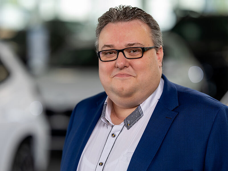 Kaltenbach Gruppe – Ein Mann mit kurzem, grauem Haar und Brille, der eine blaue Anzugsjacke und ein weißes Hemd trägt, steht in einem Innenraum mit einem unscharfen Hintergrund aus weißen Fahrzeugen in Kaltenbach Waldbröl. – BMW, MINI, KIA, BMW Motorrad & Honda Motorrad Partner in NRW