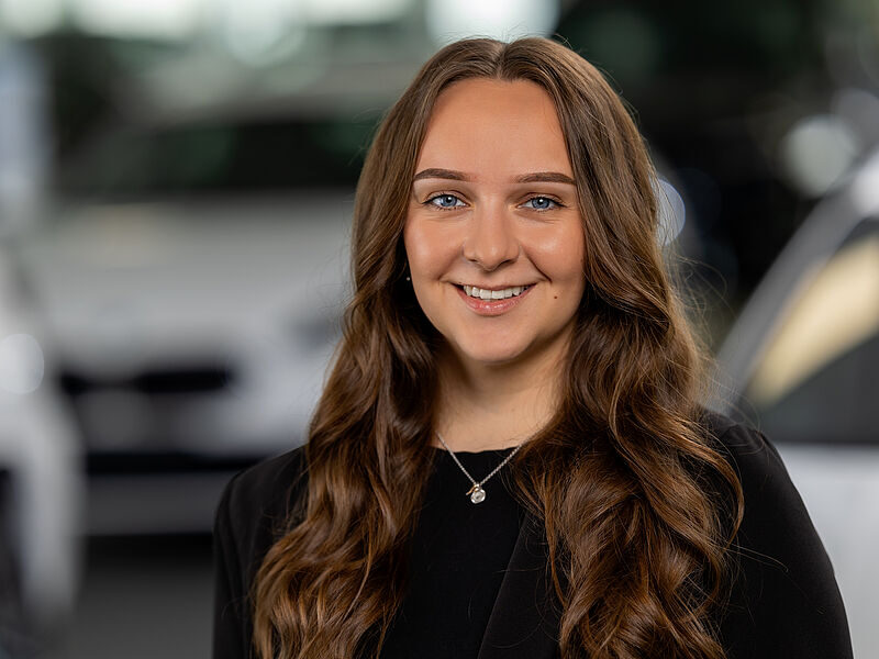 Kaltenbach Gruppe – Eine junge Frau mit langen, gewellten braunen Haaren lächelt in die Kamera. Sie trägt ein schwarzes Oberteil und eine silberne Halskette. Der unscharfe Hintergrund deutet auf eine Ausstellungshalle hin, möglicherweise in Kaltenbach-Wiehl, wobei hinter ihr Autos zu sehen sind. – BMW, MINI, KIA, BMW Motorrad & Honda Motorrad Partner in NRW