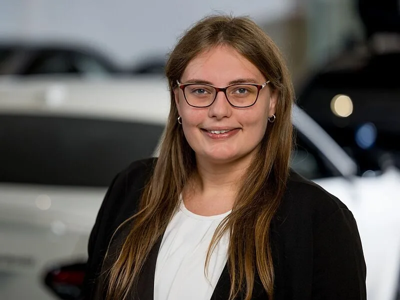 Kaltenbach Gruppe – Eine Frau mit langen braunen Haaren und Brille, die einen schwarzen Blazer und ein weißes Oberteil trägt, lächelt in die Kamera. Sie steht in einem Innenraum in Kaltenbach Wipperfürth mit unscharfen Autos im Hintergrund. – BMW, MINI, KIA, BMW Motorrad & Honda Motorrad Partner in NRW