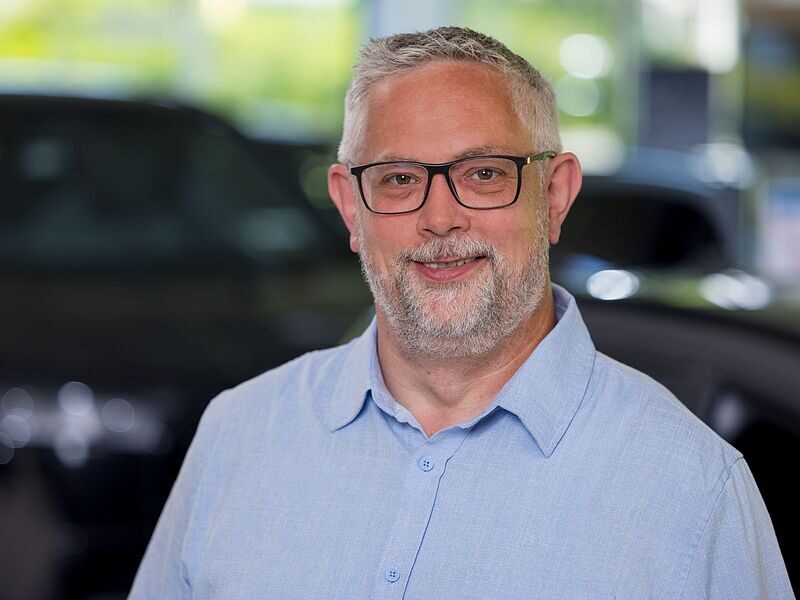 Kaltenbach Gruppe – Ein Mann mittleren Alters mit kurzen grauen Haaren, Brille und Bart lächelt in die Kamera. Er trägt ein hellblaues Button-up-Hemd und steht in einem Innenraum in Kaltenbach Overath, mit verschwommenen Autos und Fenstern im Hintergrund. – BMW, MINI, KIA, BMW Motorrad & Honda Motorrad Partner in NRW