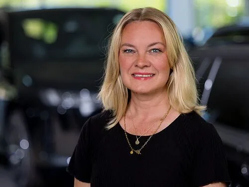 Kaltenbach Gruppe – Eine lächelnde Frau mit blonden Haaren, die ein schwarzes Oberteil und goldene Halsketten trägt, steht in einem Haus in Kaltenbach Meinerzhagen, mit unscharfen Autos im Hintergrund. – BMW, MINI, KIA, BMW Motorrad & Honda Motorrad Partner in NRW