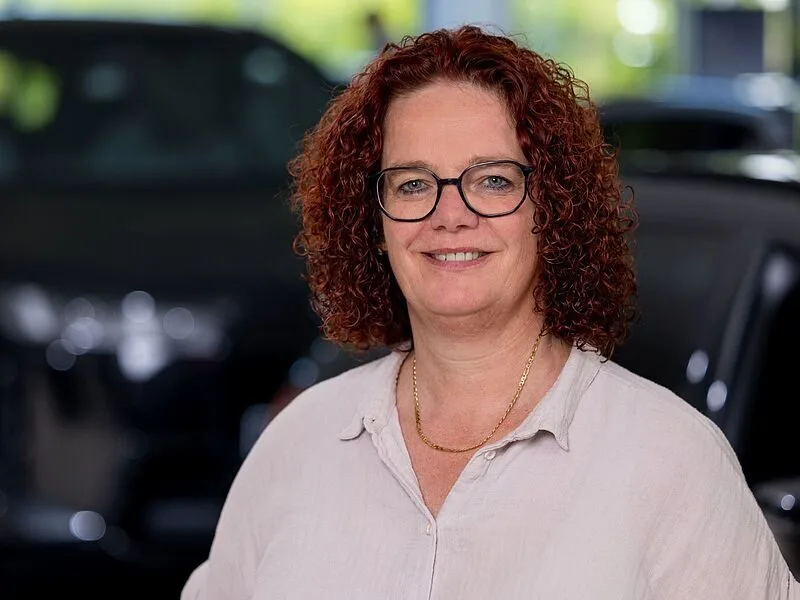 Kaltenbach Gruppe – Eine Frau mit roten Locken und Brille, die eine helle Bluse und eine goldene Halskette trägt, lächelt, während sie vor einem unscharfen Hintergrund mit einem dunkelfarbigen Auto in Kaltenbach Wiehl steht. – BMW, MINI, KIA, BMW Motorrad & Honda Motorrad Partner in NRW
