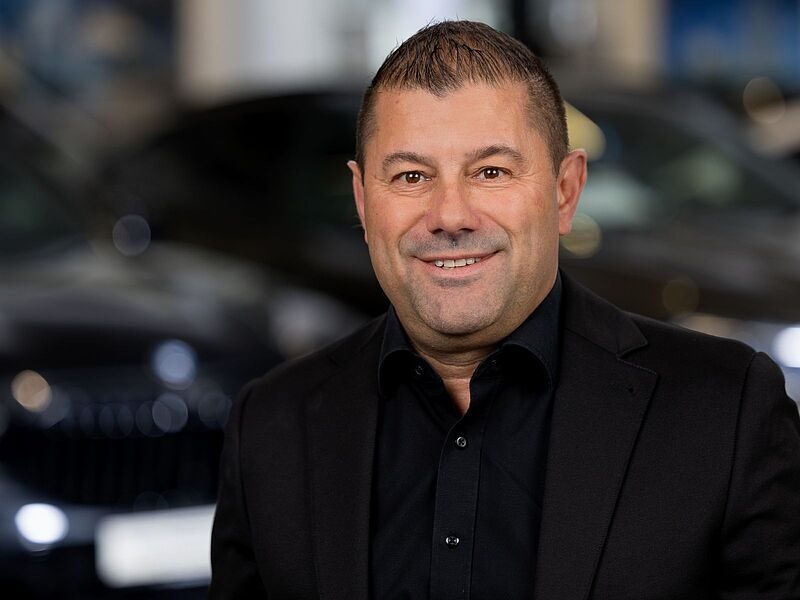 Kaltenbach Gruppe – Ein Mann in schwarzem Anzug und Hemd lächelt in die Kamera. Er steht in einem Haus in Kaltenbach-Remscheid, im Hintergrund sind unscharfe dunkle Autos zu sehen. – BMW, MINI, KIA, BMW Motorrad & Honda Motorrad Partner in NRW