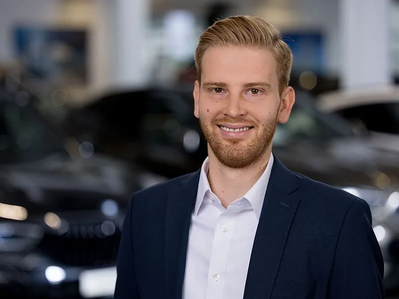 Kaltenbach Gruppe – Ein junger Mann mit hellem Haar und Bart, der ein weißes Hemd und einen dunklen Blazer trägt, steht lächelnd vor verschwommenen Autos im Autohaus Kaltenbach in Wermelskirchen. – BMW, MINI, KIA, BMW Motorrad & Honda Motorrad Partner in NRW
