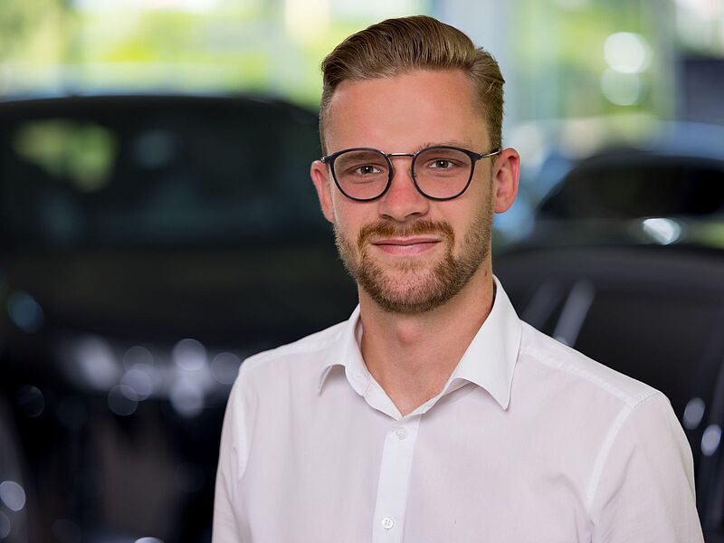 Kaltenbach Gruppe – Ein junger Mann mit kurzem, hellbraunem Haar, Bart und Brille lächelt, während er ein weißes Hemd trägt. Der unscharfe Hintergrund deutet auf einen Innenbereich hin, möglicherweise das Büro oder den Ausstellungsraum von Kaltenbach Overath. – BMW, MINI, KIA, BMW Motorrad & Honda Motorrad Partner in NRW