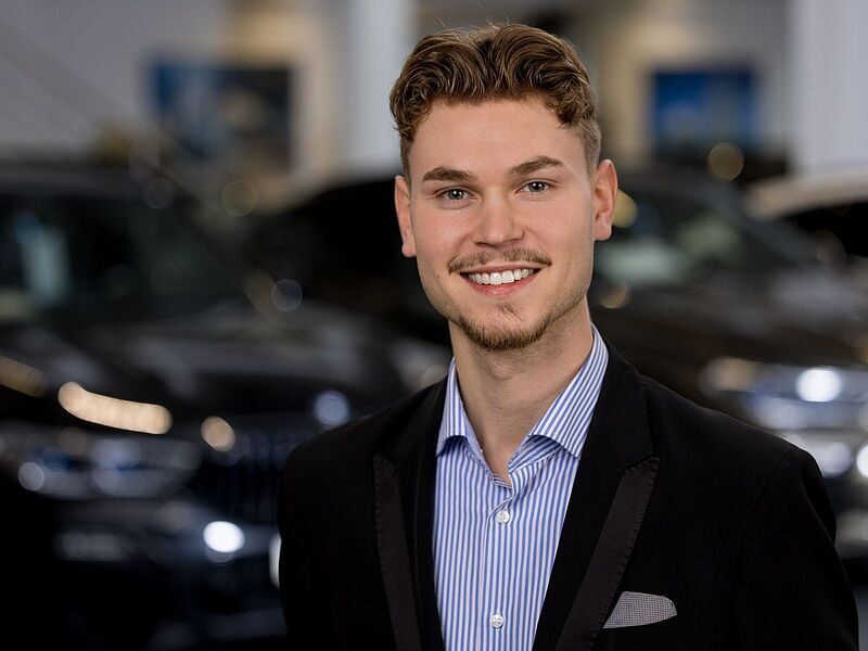 Kaltenbach Gruppe – Ein junger Mann mit kurzen, gewellten Haaren und gestutztem Bart lächelt in die Kamera. Er trägt eine dunkle Anzugjacke über einem gestreiften Hemd und steht im Ausstellungsraum von Kaltenbach Remscheid mit verschwommenen Autos im Hintergrund. – BMW, MINI, KIA, BMW Motorrad & Honda Motorrad Partner in NRW