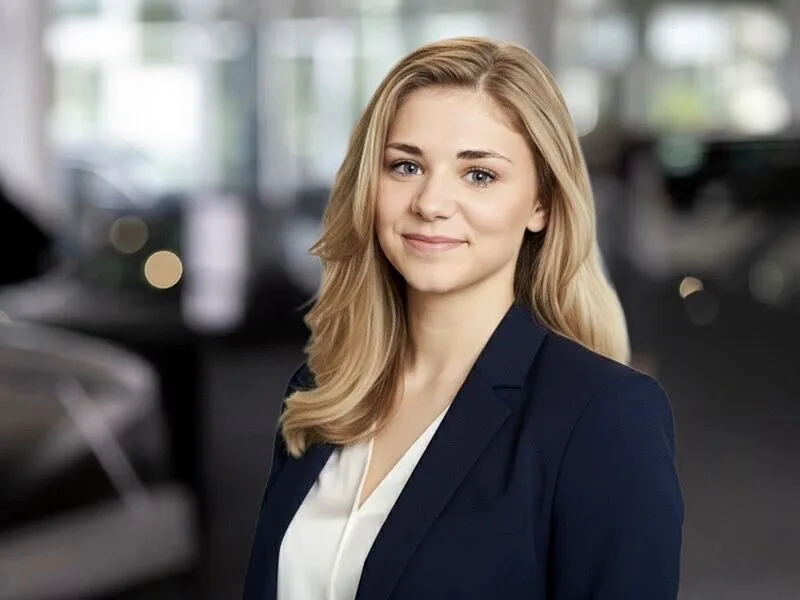 Kaltenbach Gruppe – Eine junge Frau mit langen blonden Haaren, die einen marineblauen Blazer und eine weiße Bluse trägt, lächelt sanft in die Kamera. Der verschwommene Hintergrund deutet auf ein modernes Büro hin, in dem sie als vertrauenswürdige Ansprechpartnerin fungiert. – BMW, MINI, KIA, BMW Motorrad & Honda Motorrad Partner in NRW