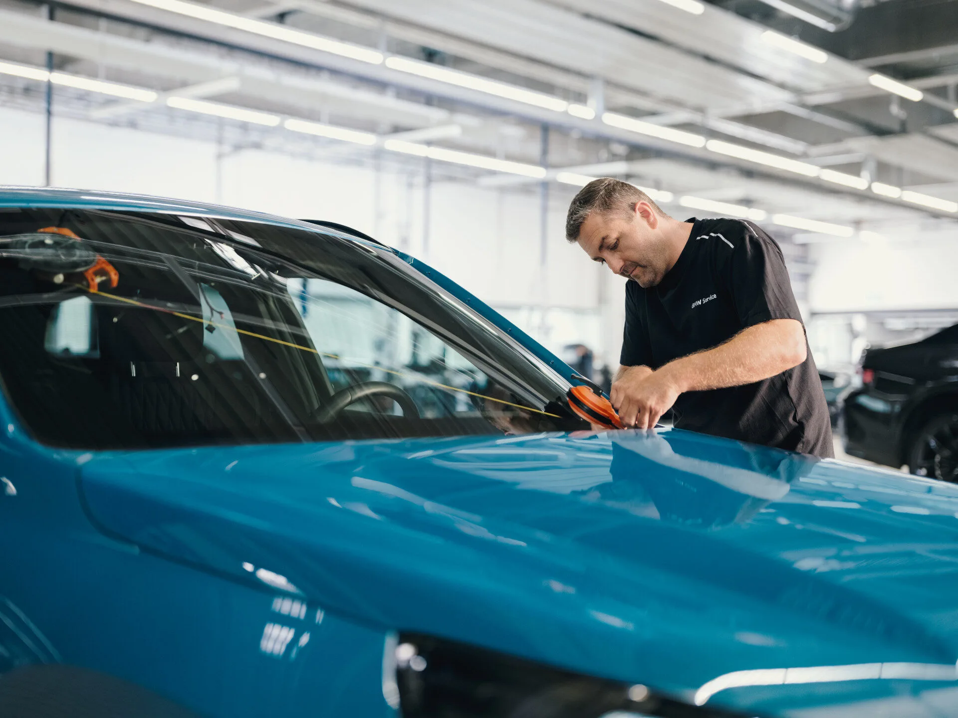 Kaltenbach Gruppe – Ein Mann in einem schwarzen Hemd konzentriert sich auf die Reparatur der Windschutzscheibe eines blauen Autos in einer hell erleuchteten Autowerkstatt, um das Glas nach einem Unfall sorgfältig zu reinigen oder zu reparieren. – BMW, MINI, KIA, BMW Motorrad & Honda Motorrad Partner in NRW