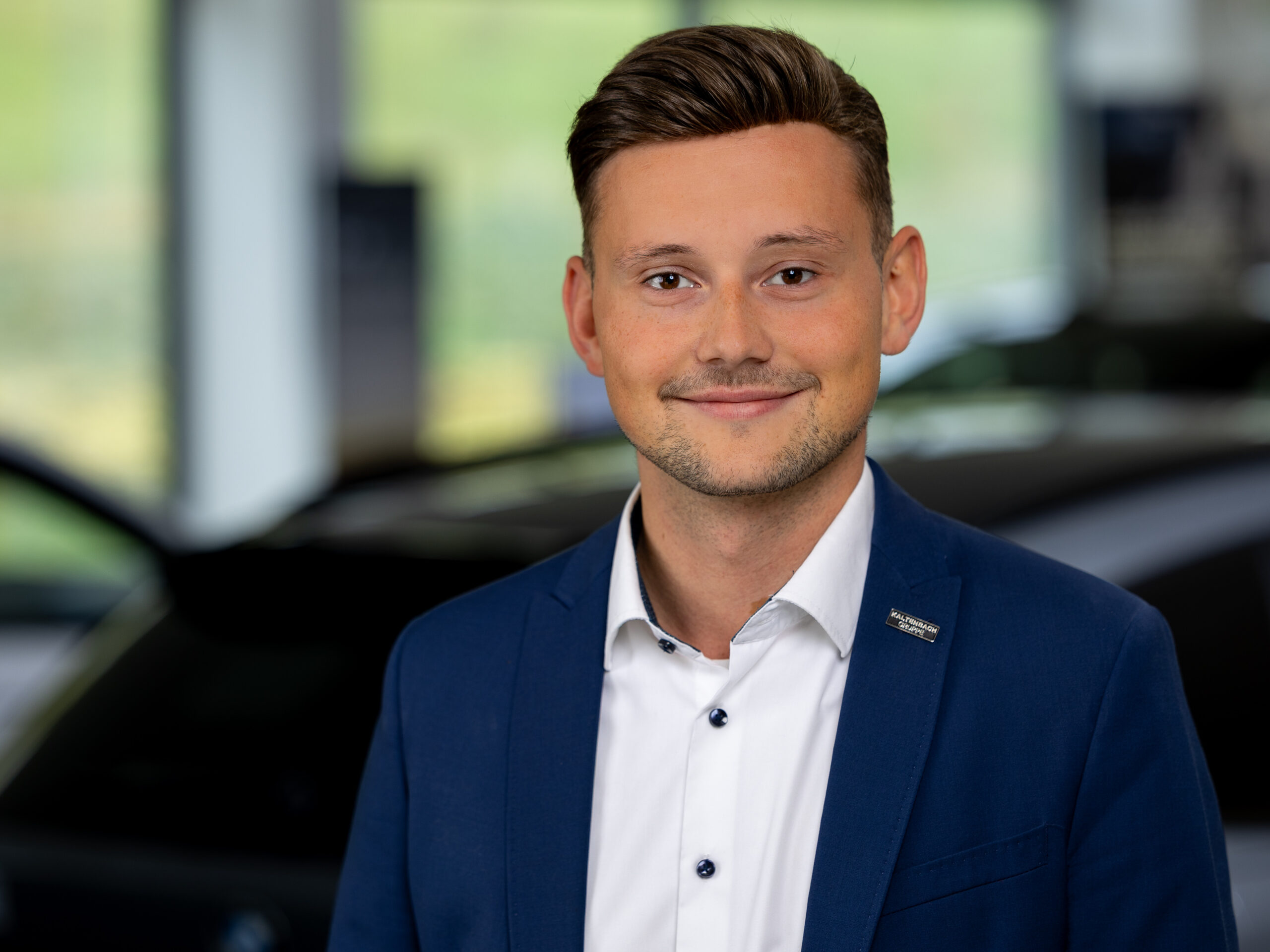 Kaltenbach Gruppe – Ein junger Mann in blauer Anzugsjacke und weißem Hemd lächelt in die Kamera. Er steht in einem Haus mit unscharfen Autos im Hintergrund in Kaltenbach Overath. – BMW, MINI, KIA, BMW Motorrad & Honda Motorrad Partner in NRW