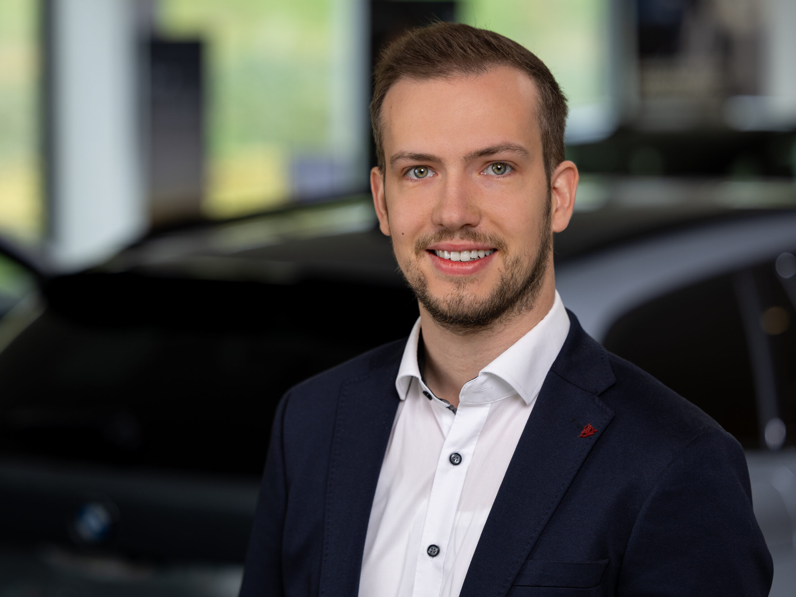 Kaltenbach Gruppe – Ein junger Mann mit kurzen Haaren und gestutztem Bart, bekleidet mit einem marineblauen Anzug und einem weißen Hemd, lächelt in die Kamera. Er steht in einem Innenraum in Kaltenbach Arnsberg, mit einem unscharfen Auto und Fenster im Hintergrund. – BMW, MINI, KIA, BMW Motorrad & Honda Motorrad Partner in NRW