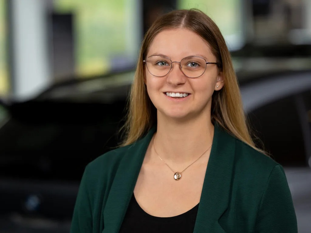 Kaltenbach Gruppe – Eine junge Frau mit langen glatten Haaren, die eine Brille, einen grünen Blazer und eine Halskette trägt, lächelt in die Kamera. Der unscharfe Hintergrund zeigt ein Auto in einer Halle in Kaltenbach-Remscheid. – BMW, MINI, KIA, BMW Motorrad & Honda Motorrad Partner in NRW
