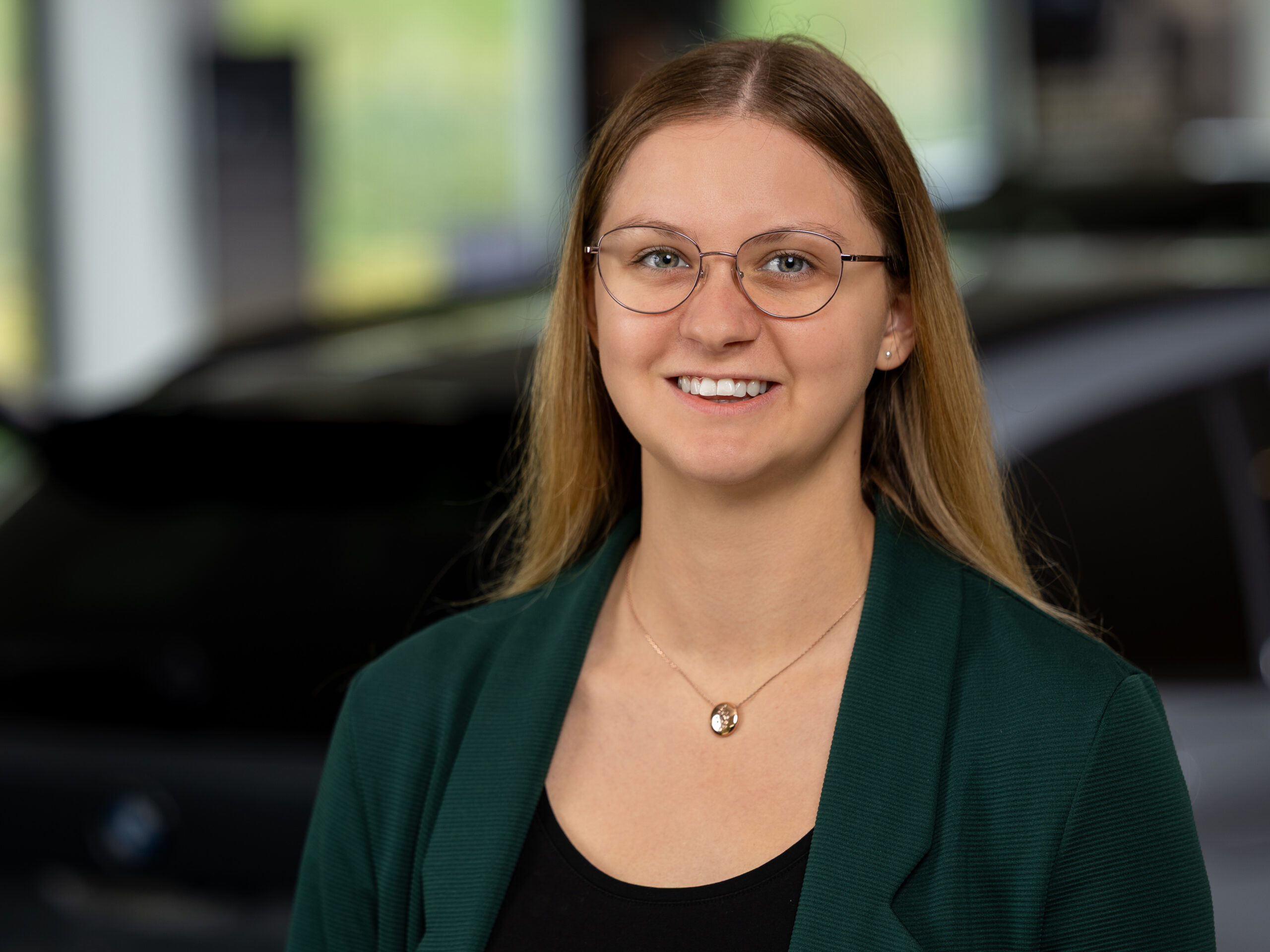 Kaltenbach Gruppe – Eine junge Frau mit langen glatten Haaren, die eine Brille, einen grünen Blazer und eine Halskette trägt, lächelt in die Kamera. Der unscharfe Hintergrund zeigt ein Auto in einer Halle in Kaltenbach-Remscheid. – BMW, MINI, KIA, BMW Motorrad & Honda Motorrad Partner in NRW
