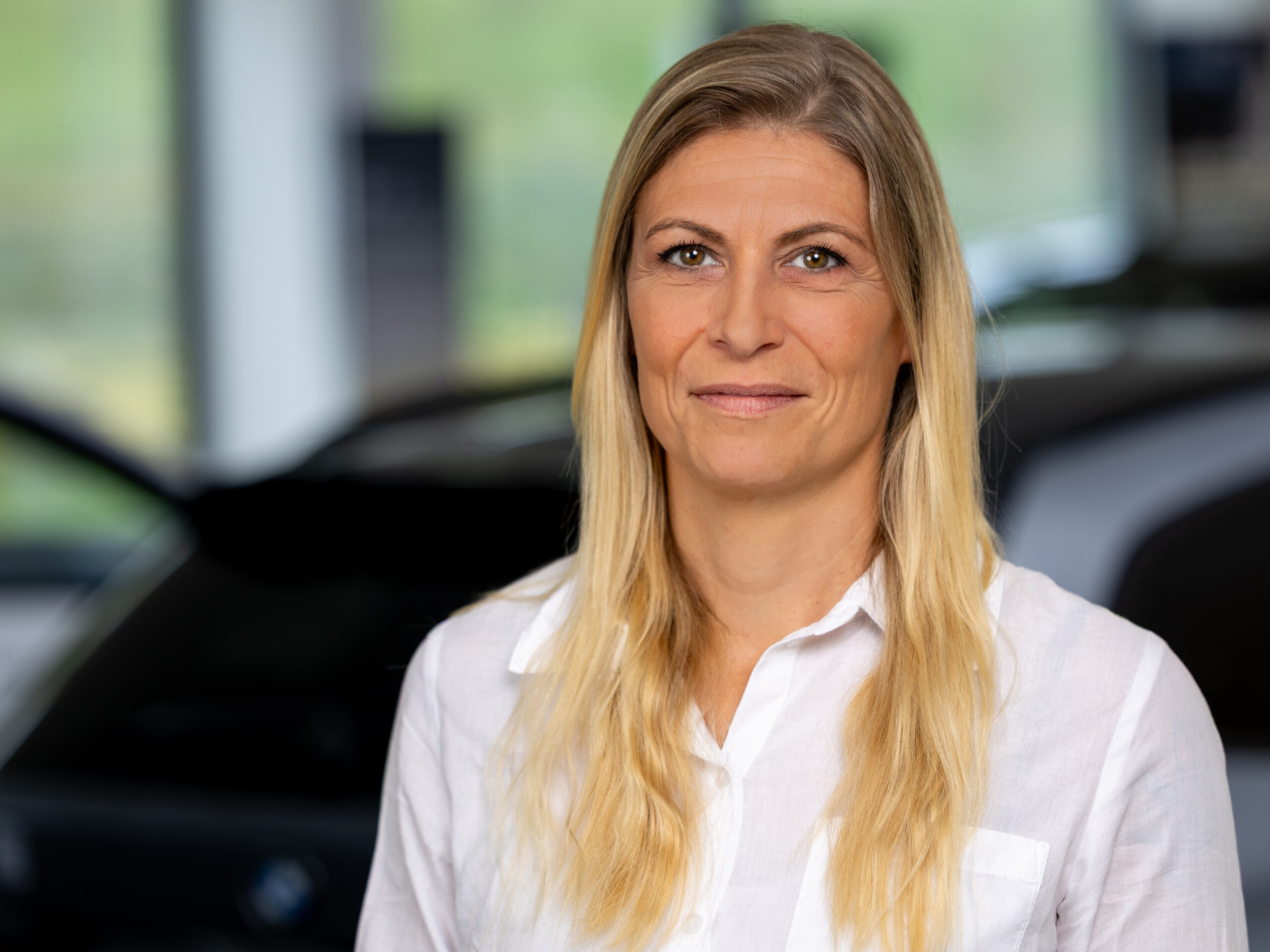 Kaltenbach Gruppe – Eine Frau mit langen blonden Haaren und weißem Hemd steht in einem Innenraum, wahrscheinlich im Ausstellungsraum von Kaltenbach Remscheid, vor einem unscharfen Hintergrund mit Autos. Sie blickt in die Kamera und lächelt leicht. – BMW, MINI, KIA, BMW Motorrad & Honda Motorrad Partner in NRW